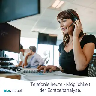 Mitarbeiterin führt ein Telefongespräch in einem modernen Büro mit Kollegen im Hintergrund.